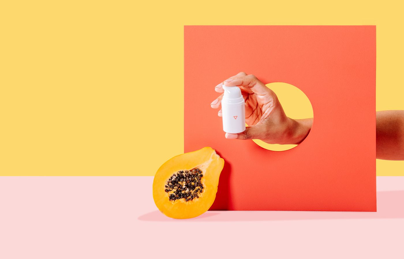 Hand holding a bottle of Wisp Estradiol Cream on pink and yellow background with papaya fruit