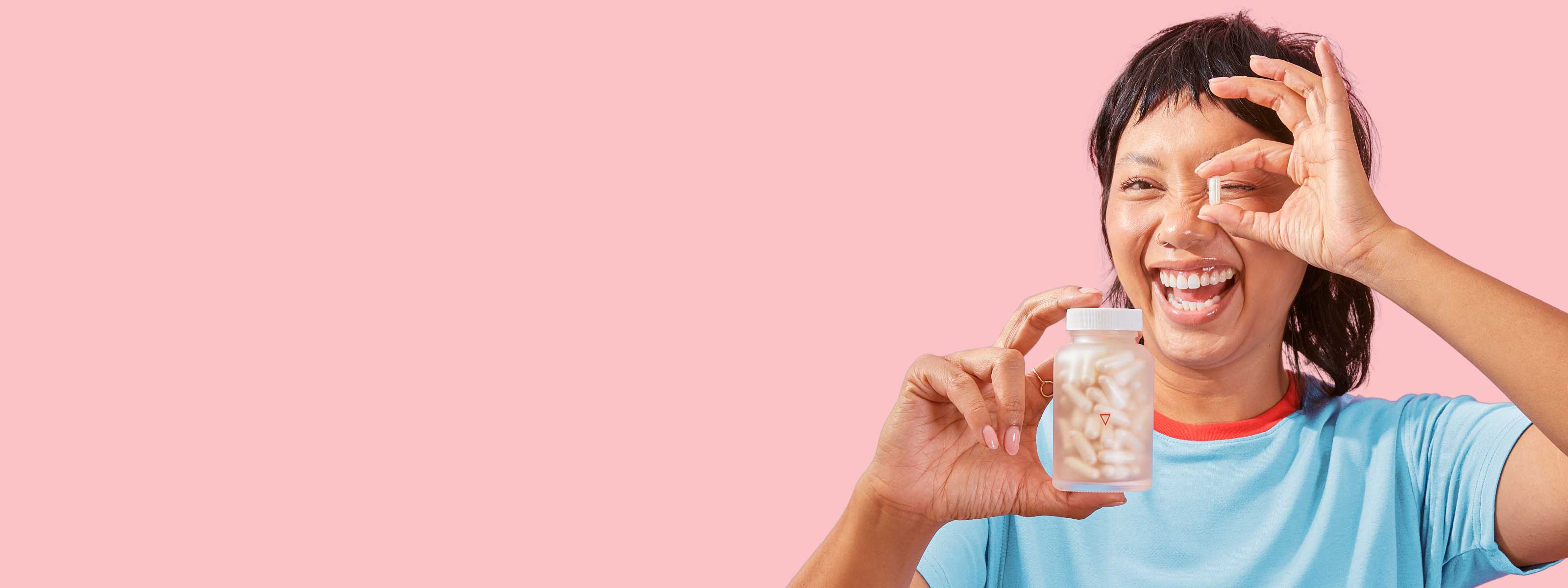 A smiling female holding a Wisp glass pill jar in one hand and a a pill in the other in front of a pink background