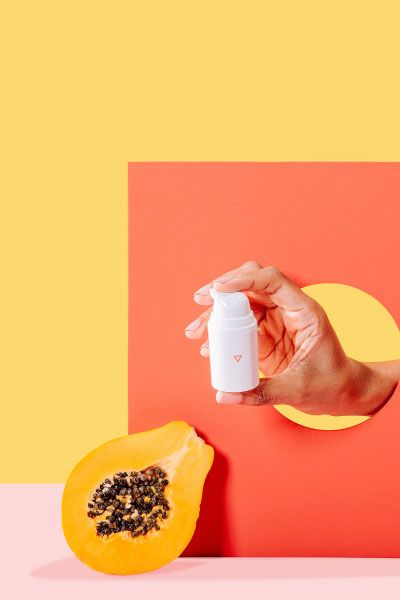 A woman's hand holding a bottle of Estradiol Cream with a papaya on a yellow and pink background