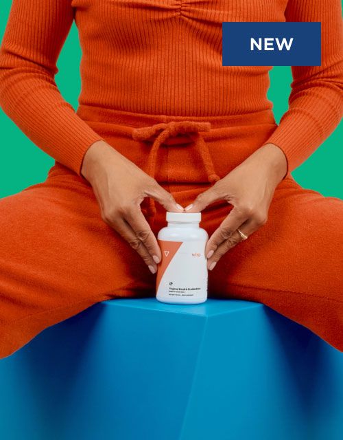 A woman wearing a red jumpsuit is sitting on a blue block with a bottle of Wisp Vaginal Probiotics in front of her