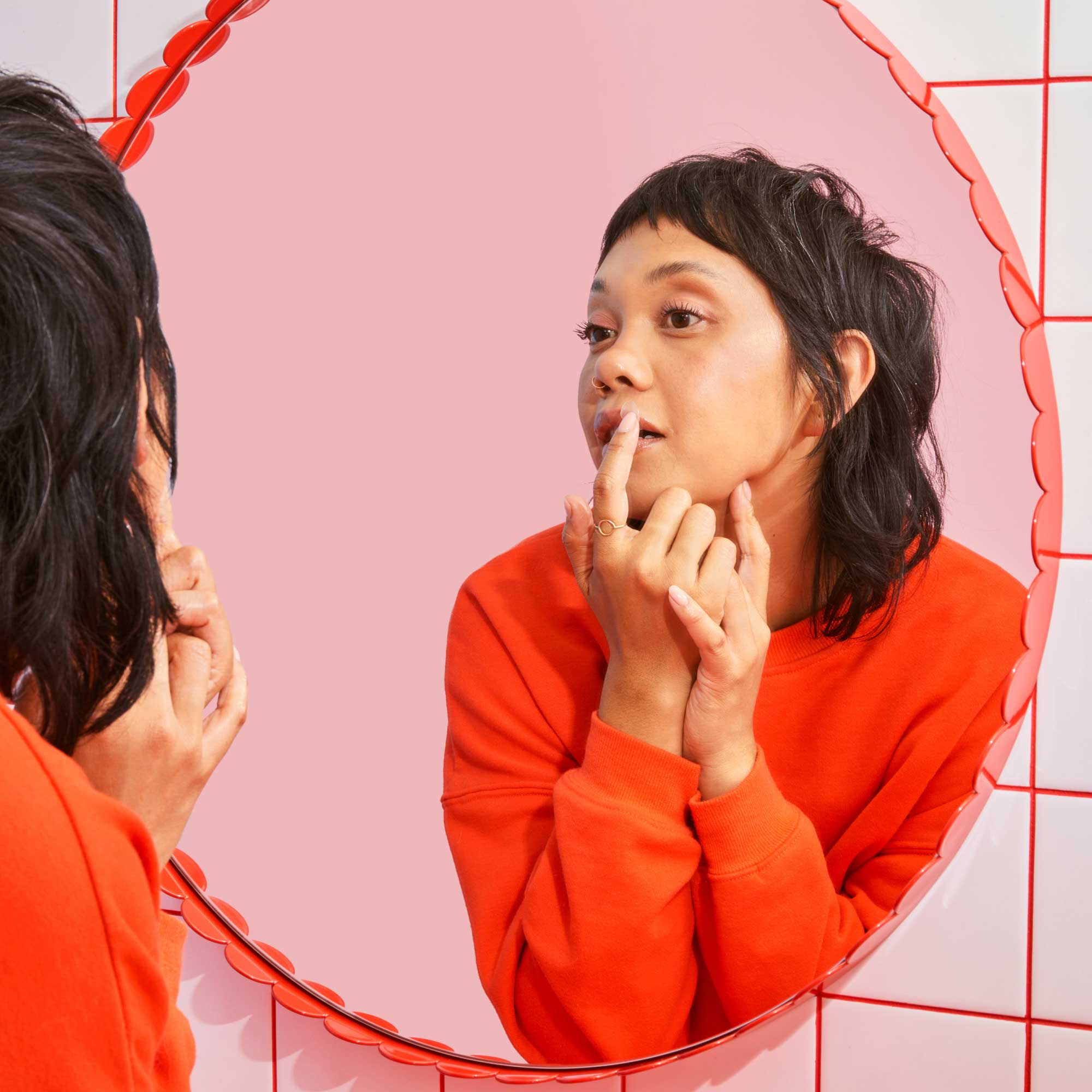 A woman wearing a red sweater looking in the mirror to put a hydrocolloid patch on a cold sore on her lip