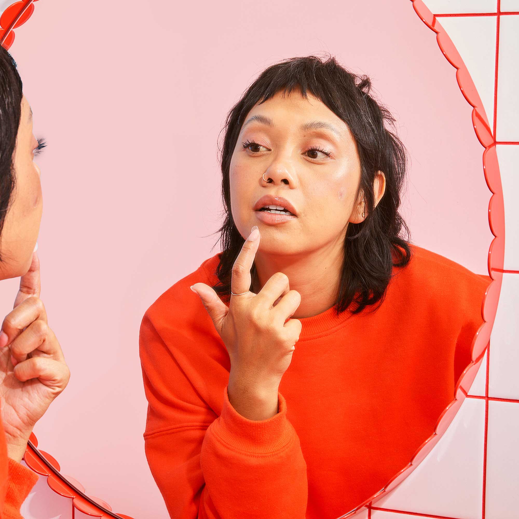 A woman wearing a red sweater looking in the mirror to put a hydrocolloid patch on a pimple on her chin