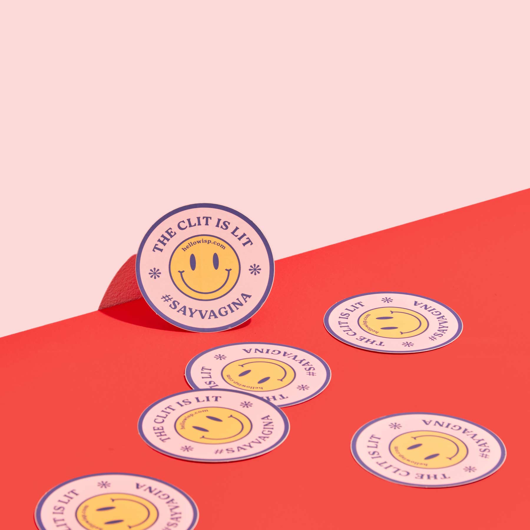 Wisp stickers with a pink background and yellow smiley face that say the clit is lit