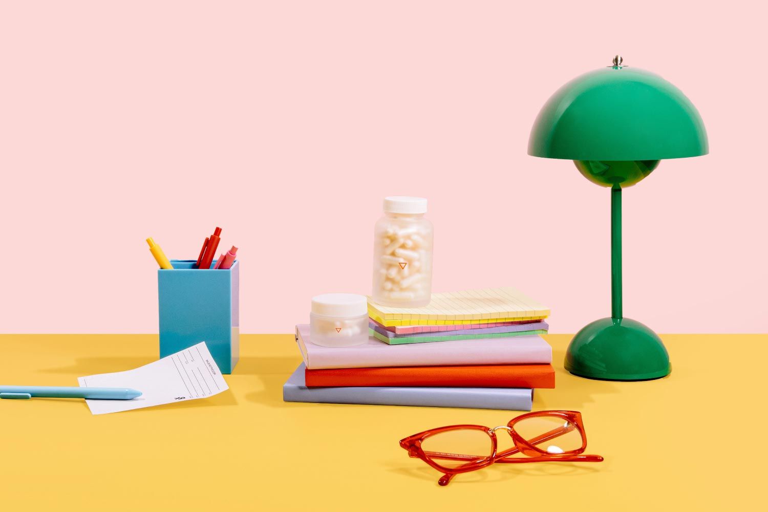 Wisp glass jars, a green lamp, pen holder, red glasses and an Rx script on a yellow and pink background