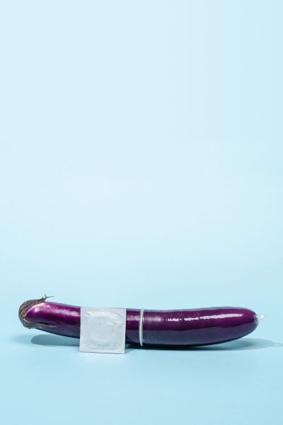 An eggplant with a condom on it and a silver unopened condom wrapper on a light blue surface