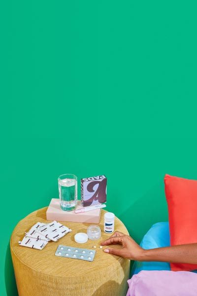 A woman's hand reaching for pills on a yellow nightstand with Wisp's medication abortion kit in front of a green wall with red, blue and pink bedding