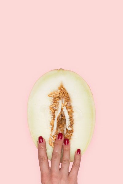 A woman's hand touching a vulva-shaped cut open fruit on a pink surface