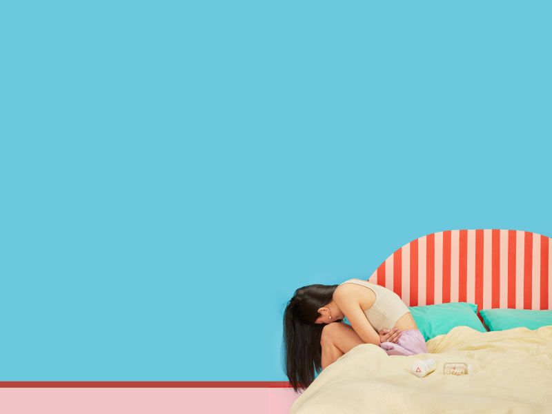 A woman wearing a tank top and shorts is sitting on a colorful bed doubled over in discomfort