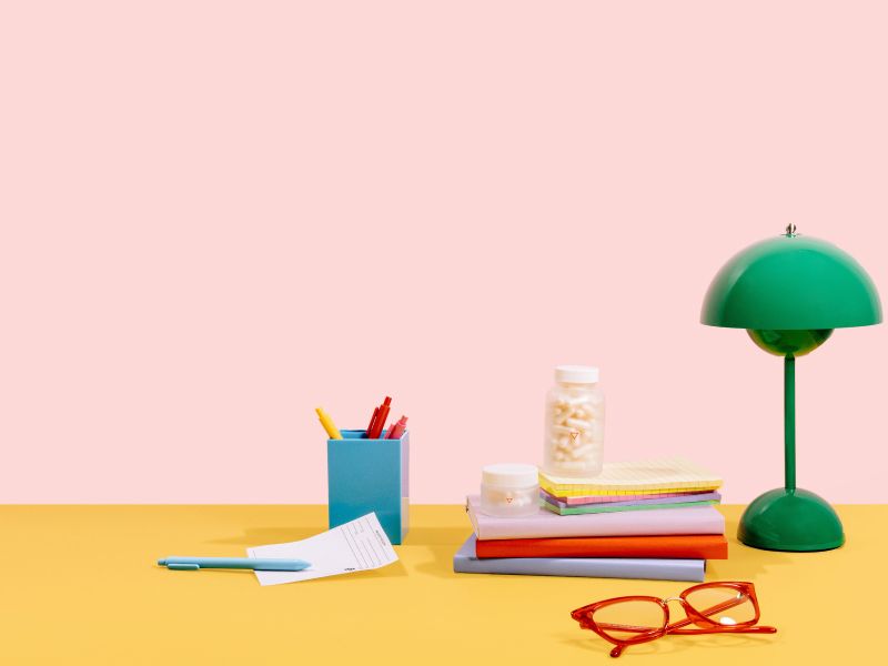 A desktop scene with a green lamp, red glasses, a stack of notebooks and a blue pen holder with an rx script and two Wisp glass jars on a pink and yellow background