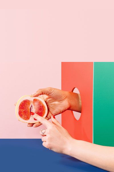 Woman's hand holding a grapefruit with colorful abstract shapes on a pink and blue background
