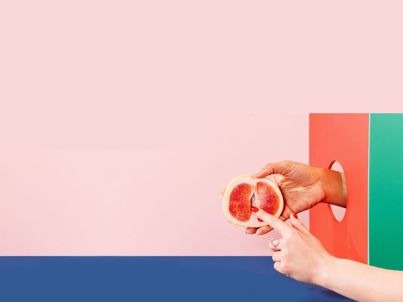 Woman's hand holding a grapefruit with colorful abstract shapes on a pink and blue background