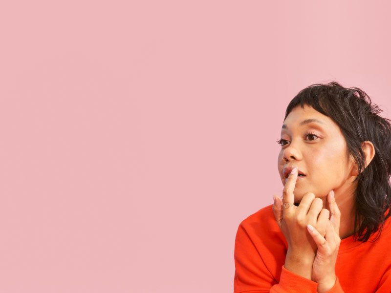 A female with dark hair and wearing a red sweater is looking in the mirror while putting a hydrocolloid patch on a cold sore