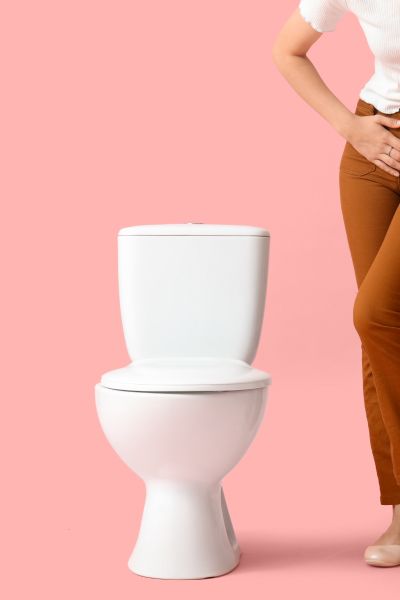 A woman standing next to a toilet wearing a white shirt and brown pants with her hands on her pelvic region indicating discomfort