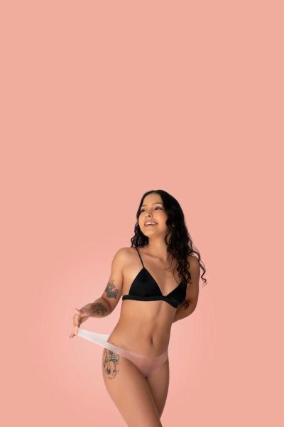 A female wearing a black bra and latex Lorals underwear in front of a peach background