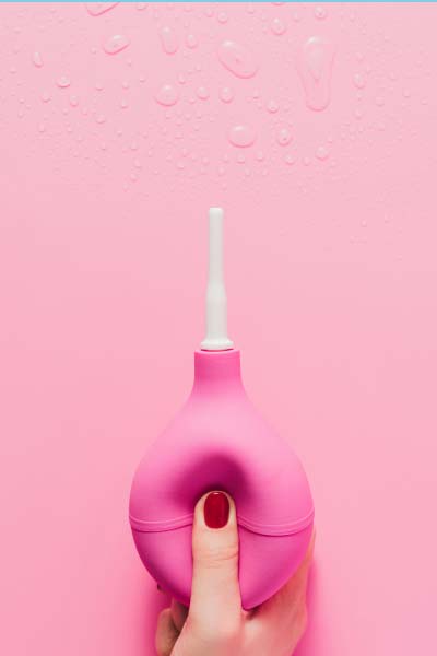 A woman's hand with red fingernails holding a pink vaginal douche bulb over a pink background