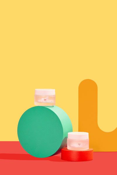 Two small Wisp glass pill jars balanced on colorful abstract shapes on a red surface with a yellow background