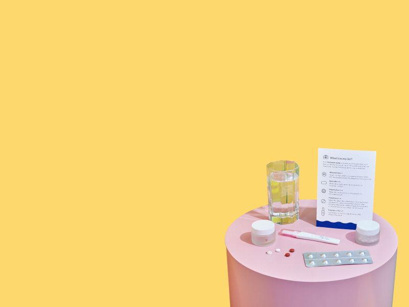 A pink nightstand with Wisp online abortion medications, treatment guide, and a glass of water, with yellow and green walls