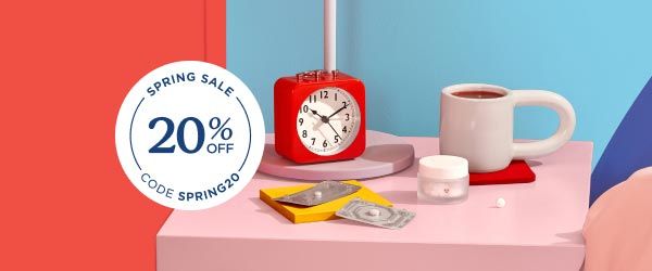 A Wisp glass pill jar and 2 emergency contraception packets are on a pink nightstand with a red alarm clock and white mug next to a bed with colorful bedding