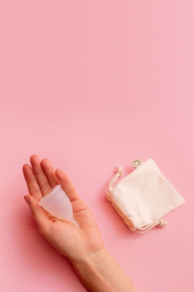 A woman's hand holding a menstrual cup with a storage bag next to it on a pink surface