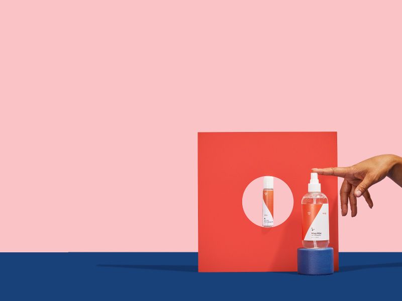 a woman's hand touches Wisp Mist adult toy cleaner next to Toy-Safe Harmonizing Lube on a colorful geometric background