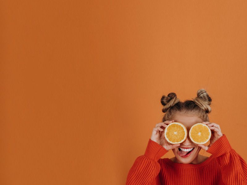 a woman in a red sweater sticking her tongue out and holding orange slices over her eyes, on an orange background