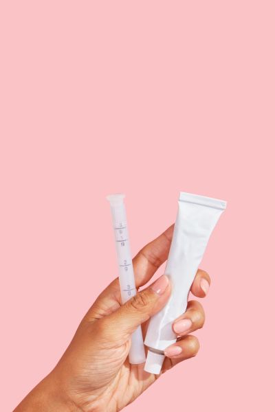 Female hand holding a tube of vaginal dryness cream and applicator with a pink background