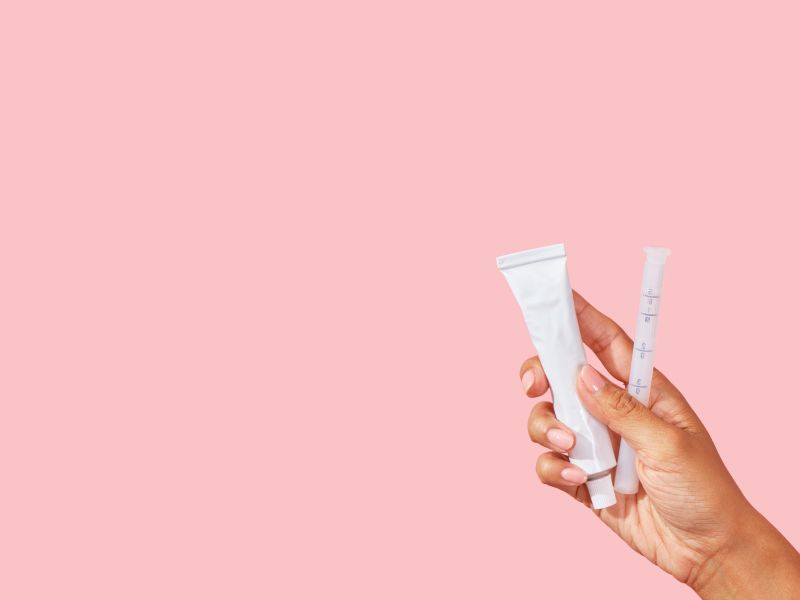 Female hand holding a tube of vaginal dryness cream and applicator with a pink background