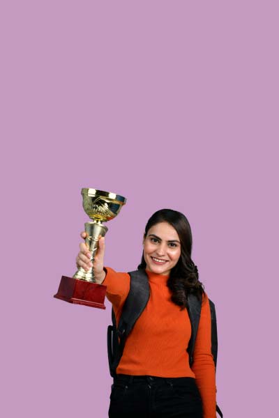 A woman wearing a red sweater, black pants and a black backpack holding a gold trophy in the air in front of a purple background