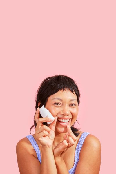 a smiling woman holding a bottle of Wisp herpes medication and applying it to her lip against a pink background