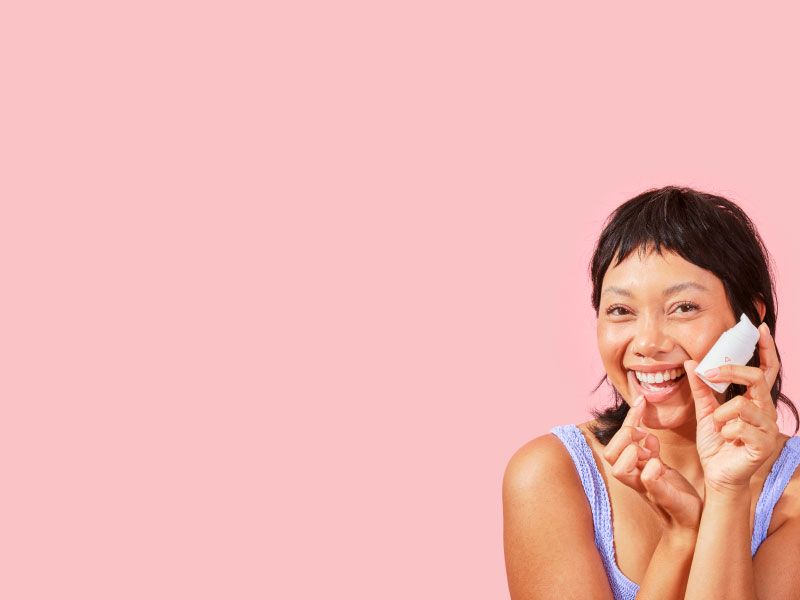 a smiling woman holding a bottle of Wisp herpes medication and applying it to her lip against a pink background