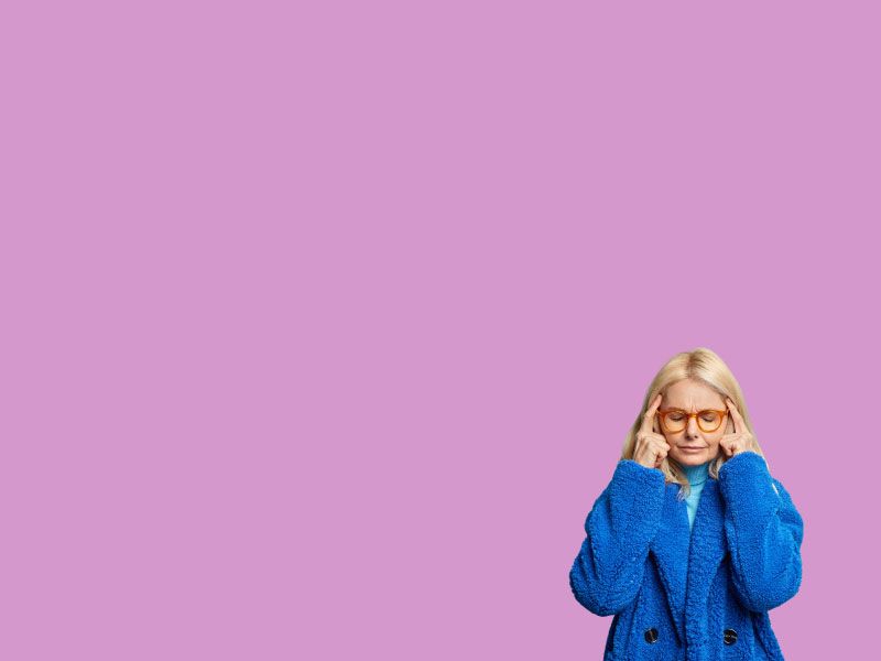 A woman with blonde hair is wearing a fluffy royal blue coat with her fingers on her temples in discomfort in front of a purple background