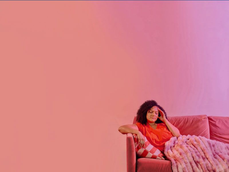 A woman wearing a red shirt is sitting on a fuschia sofa with a pink blanket and she has her hand on her temple in discomfort