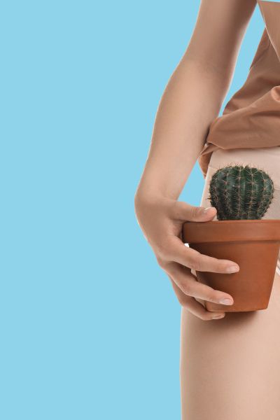 A woman standing up in white underwear and a tan shirt with a hand on her abdomen and holding a small cactus indicating pain