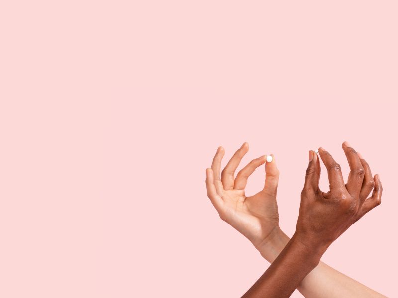 Two female hands criss crossing, both holding an Emergency Contraception pill in front of a pink background