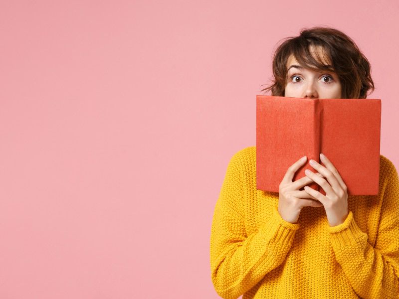 A woman wearing a yellow sweater and holding a brown book with a surprised look on her face
