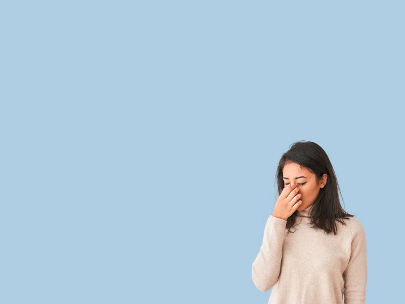 A woman wearing a cream shirt is pinching the bridge of her nose in discomfort