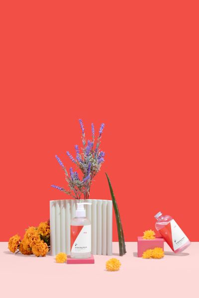 Two bottles of Balancing Wash balanced on colorful abstract shapes with lavender, Aloe Vera and Marigold on a red and pink background