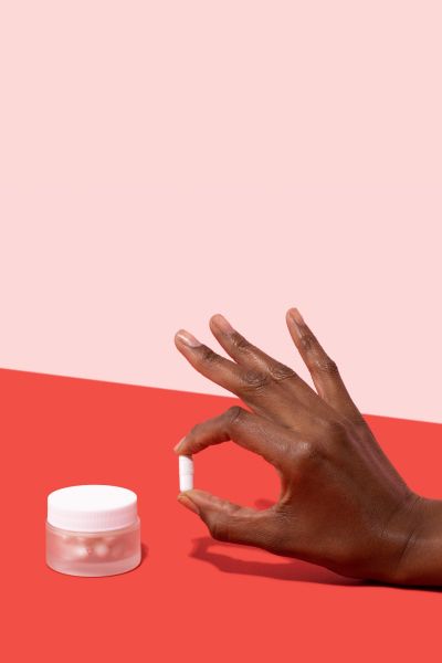 Woman's hand holding a Boric Acid pill next to a small glass Wisp jar on a pink and red background