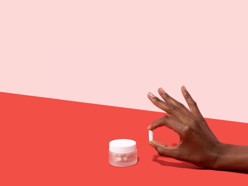 Woman's hand holding a Boric Acid pill next to a small glass Wisp jar on a pink and red background