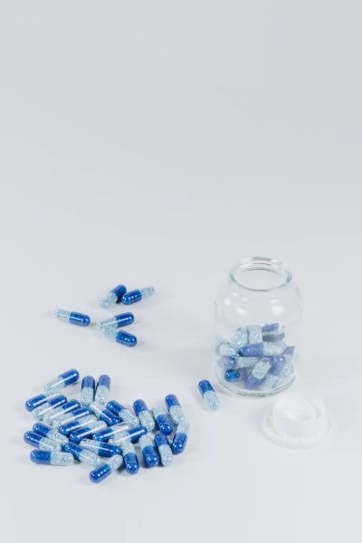 A clear pill bottle with blue and white pills inside and scattered around the bottle