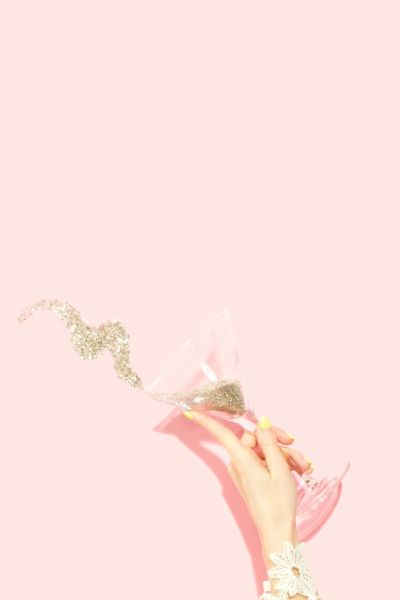 a woman's hand holding a martini glass pouring glitter onto a pink background