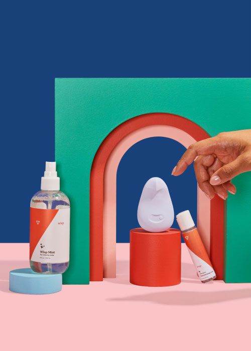 Woman's hand reaching for Dame Pom, Toy-Safe Lube, and Wisp Mist Toy Cleaner with colorful abstract shapes on a blue and pink background