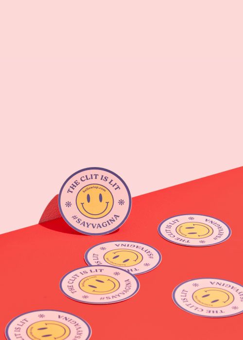 Pink stickers with a smiley face that say the clit is lit sitting on a red surface against a pink background