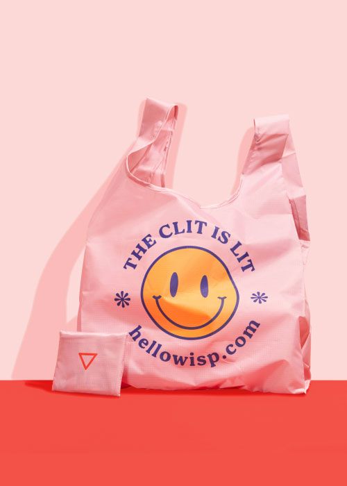 Pink foldable shopping tote with a smiley face that says the clit is lit sitting on a red surface against a pink background
