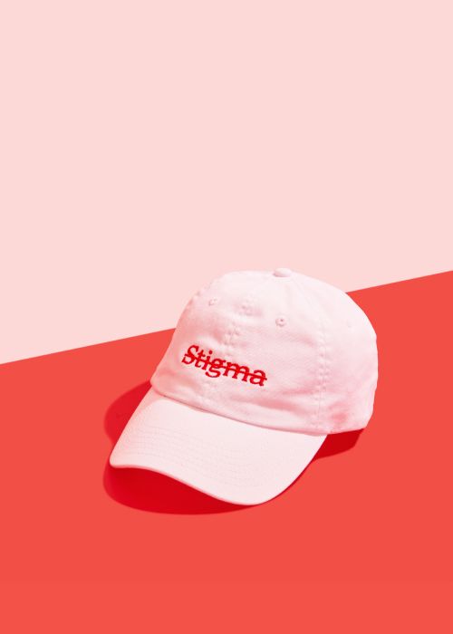 White baseball cap with red Stigma text crossed out on front and red Wisp logo on back, sitting on a red surface on a pink background