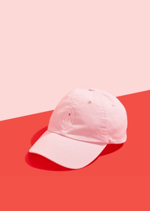 Pink baseball cap with white and red vulva design on front and red Wisp logo on back, sitting on a red surface on a pink background