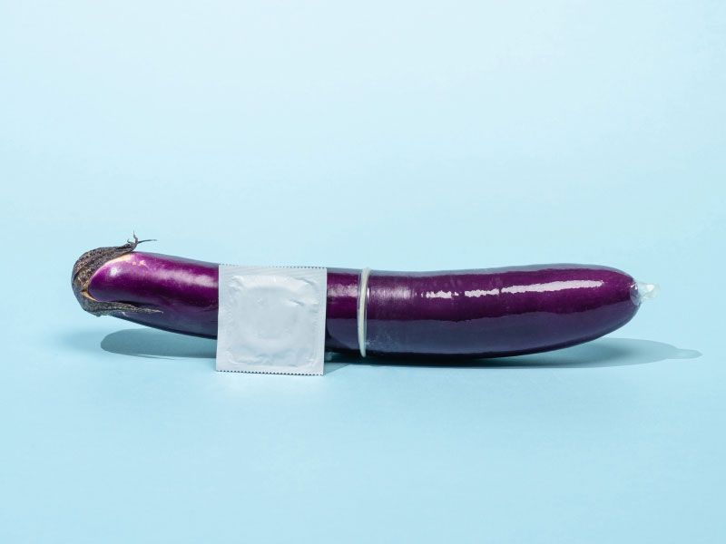 An eggplant with a condom on it and a silver unopened condom wrapper on a light blue surface