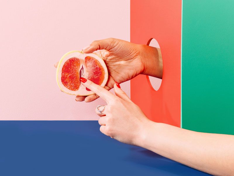 Woman's hand holding a grapefruit with colorful abstract shapes on a pink and blue background
