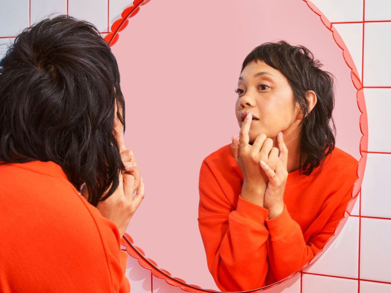 A female with dark hair and wearing a red sweater is looking in the mirror while putting a hydrocolloid patch on a cold sore