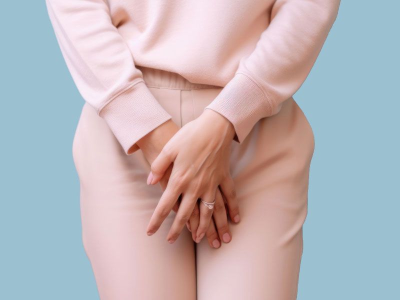 A woman wearing light pink sweater and pants with her hands over her groin indicating discomfort